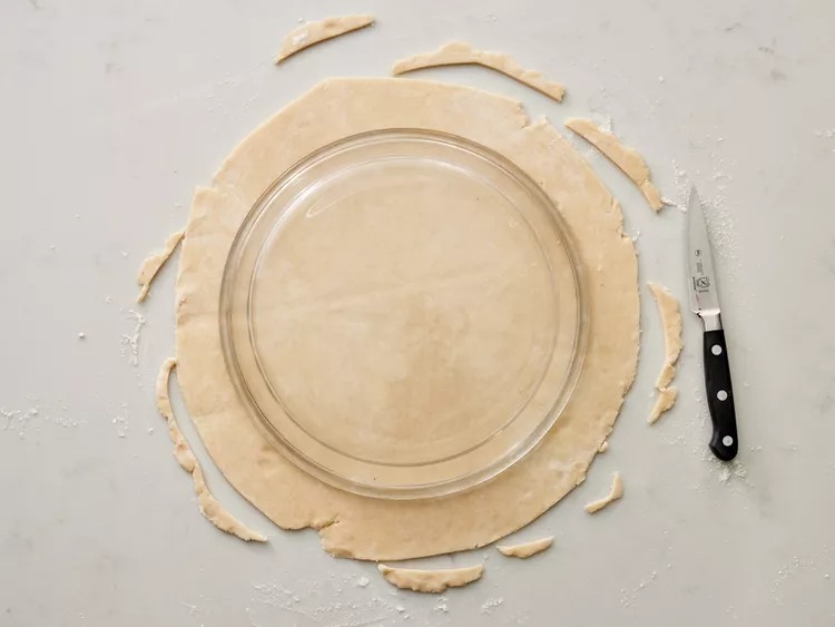 I shape the dough into a ball, dust the counter with flour, and roll it out to about 1/8 inch thick. Not perfect. Just flat enough. I flip a 9-inch pie pan upside down on the dough and cut a circle that’s about an inch and a half bigger than the pan.